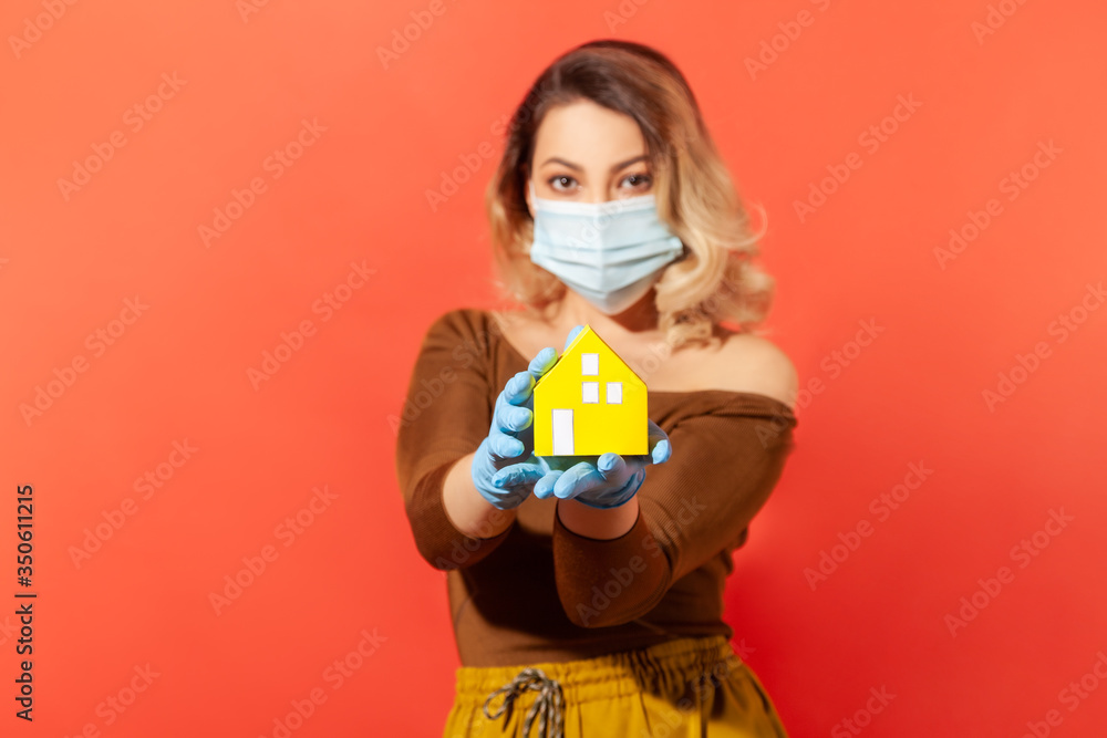 Woman in protective mask and gloves showing paper house on her hand, recommendation to stay home, adhere quarantine while contagious disease spread, coronavirus outbreak. indoor studio shot isolated Woman in protective mask and gloves showing paper house on her hand, recommendation to stay home, adhere quarantine while contagious disease spread, coronavirus outbreak. indoor studio shot isolated