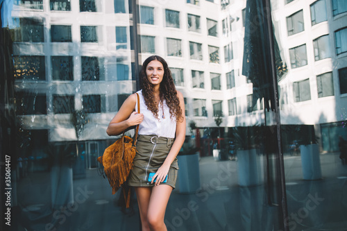 Half length portrait of smiling young 20s woman in trendy outfit with bag standing on urban setting,beautiful hipster girl looking at camera posing near glass exterior of building in town on sunny day © GalakticDreamer