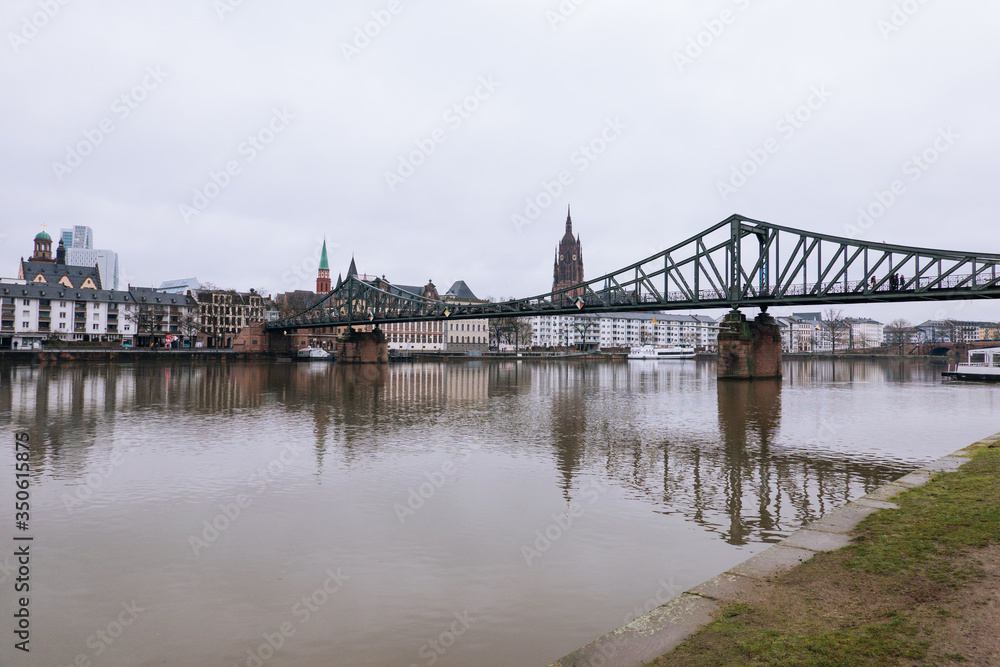 Outdoor cloudy view of Eiserner Steg, historical pedestrian Iron Bridge, and promenade on riverside of Main River with skyline in rainy day in Frankfurt, Germany.