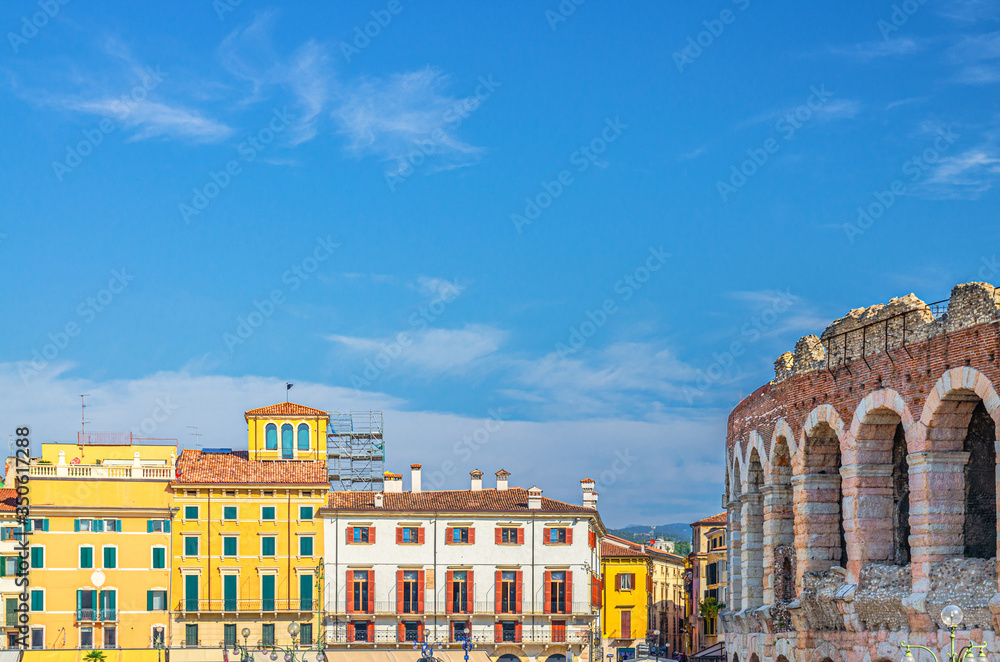 The Verona Arena limestone walls with arch windows and old colorful multicolored buildings in Piazza Bra square in Verona city historical centre, blue sky background, Veneto Region, Northern Italy
