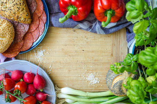 Healthy ingredients on the table. Breakfast kitchen composition with raw vegetables, salami, buns, cherry tomatoes, onions, basil, peppers, arranged in a circle on a cutting board.