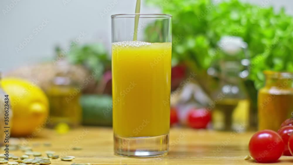 Orange juice is poured into a glass on the background of vegetables and food. Close up glass of fresh orange juice. Cooking a healthy breakfast with natural ingredients. Homemade fruit juice on the ta Orange juice is poured into a glass on the background of vegetables and food. Close up glass of fresh orange juice. Cooking a healthy breakfast with natural ingredients. Homemade fruit juice on the ta