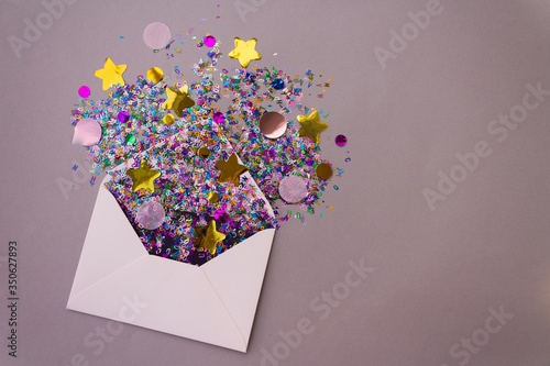 Opened white envelope with multicolored sparkle glitter confetti letters on pastel lilac background. Festive greeting concept. Flat lay style. Top view. Copy text