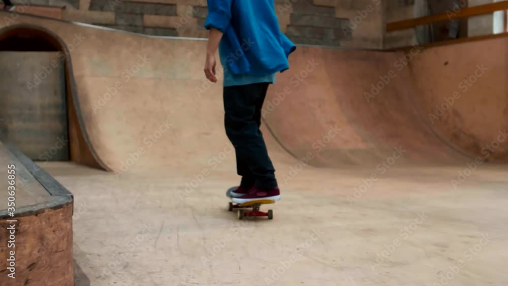 Back view of unrecognizable skater wearing blue shirt, sneakers and jeans holding his skateboard, walking along indoor skate park and starting skateboarding Back view of unrecognizable skater wearing blue shirt, sneakers and jeans holding his skateboard, walking along indoor skate park and starting skateboarding