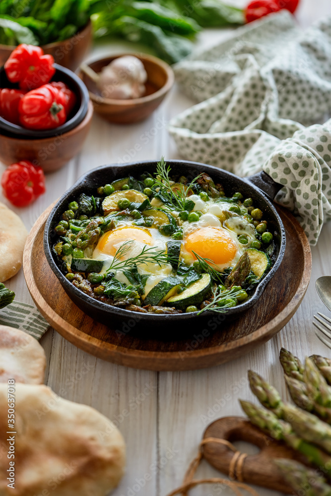 Green shakshuka with green vegetables and fried eggs in cast iron skillet on a white wooden table. Green shakshuka with green vegetables and fried eggs in cast iron skillet on a white wooden table.