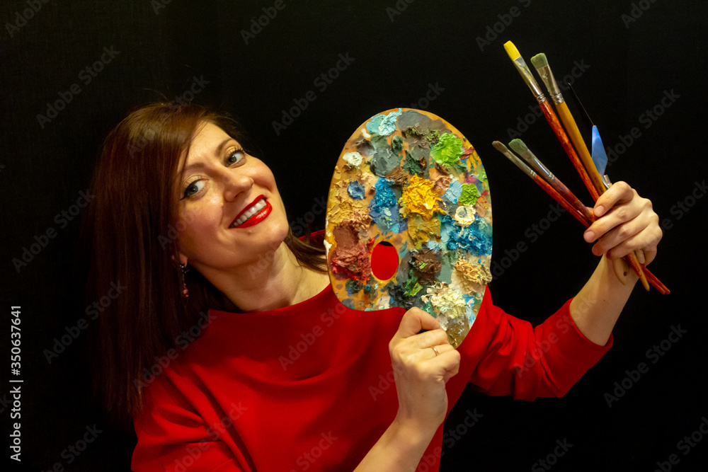 Fototapeta premium A young beautiful woman wearing red dress on black background. pretty young smiling woman holding a palette and artistic brushes.