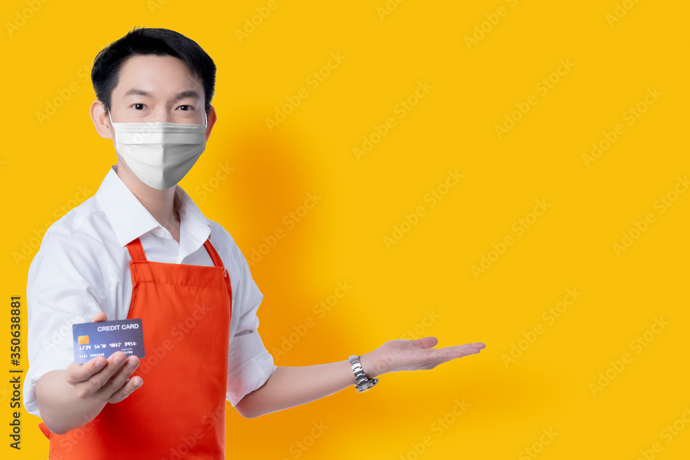 asian attractive male business owner with apron uniform and surgical mask protection hand gesture open welcome with credit card online payment method isolate yellow backgorund asian attractive male business owner with apron uniform and surgical mask protection hand gesture open welcome with credit card online payment method isolate yellow backgorund