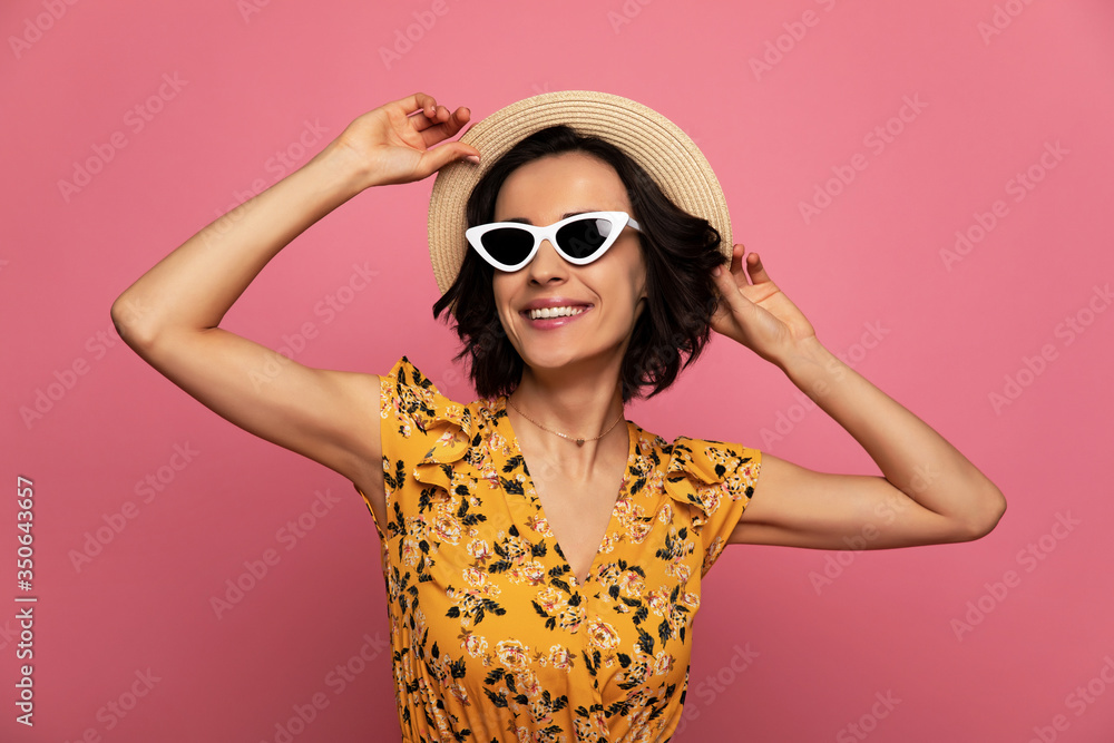 On the beach. Close-up photo of overjoyed attractive woman in a floral dress, straw hat and sunglasses, who is smiling and touching her hat. On the beach. Close-up photo of overjoyed attractive woman in a floral dress, straw hat and sunglasses, who is smiling and touching her hat.