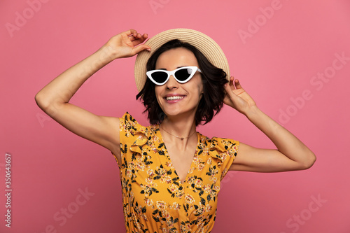 On the beach. Close-up photo of overjoyed attractive woman in a floral dress  straw hat and sunglasses  who is smiling and touching her hat.