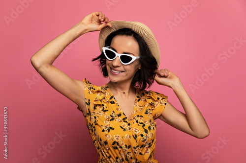 Party time. Close-up photo of a young happy woman in yellow dress and sunglasses, who is touching her straw hat and smiling.