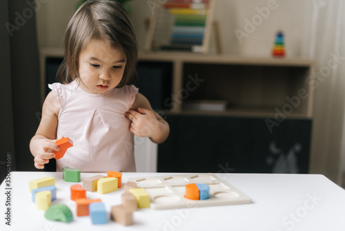 Joyful beautiful little girl is is playing with colorful wooden block toys at table in cozy children room.