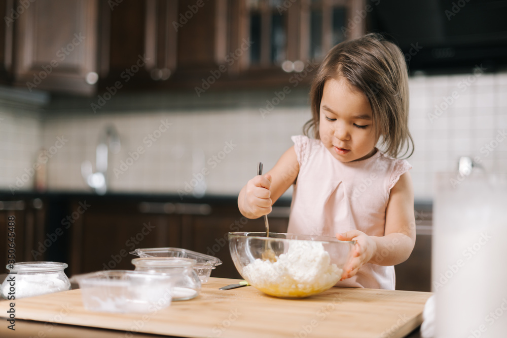 Cute beautiful little girl is kneading and mixing cottage cheese and eggs in transparent bowl for making cheesecakes at the table in kitchen with modern interior. Cute beautiful little girl is kneading and mixing cottage cheese and eggs in transparent bowl for making cheesecakes at the table in kitchen with modern interior.
