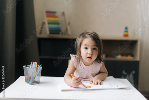 Surprised little two-year-old girl wearing casual white dress is drawing on white paper with colorful felt-tip pens at table in children's room.