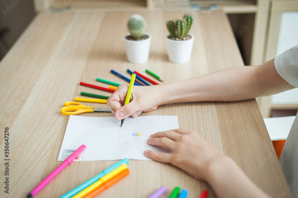 The child at the table draws on a blank sheet, around many colored pencils and felt-tip pens. Close-up of hands and pencils.