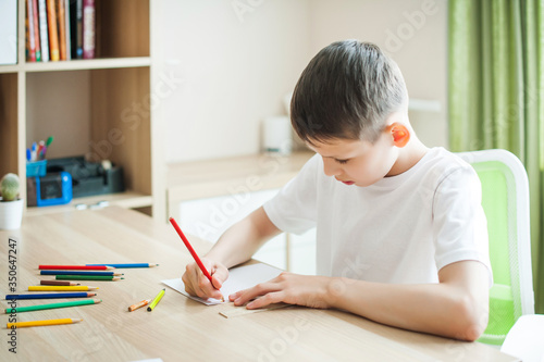 Picture of a thoughtful Caucasian schoolboy in a white T-shirt sitting on a table indoors, concentrated, draws with a pencil. On the table are pencils. Holding a pencil is not correct.