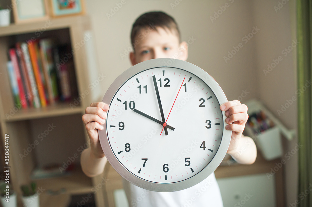 A teenager boy holds a big clock with large numbers on outstretched hands. A child in a white T-shirt, watch closeup, face in defocus. In the children's room. Concept mode, schedule, sleep. A teenager boy holds a big clock with large numbers on outstretched hands. A child in a white T-shirt, watch closeup, face in defocus. In the children's room. Concept mode, schedule, sleep.