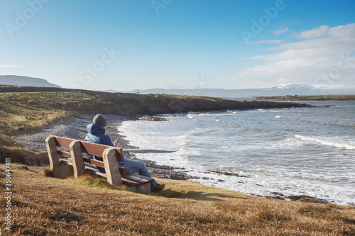 Man sitting on a bench with a beautiful view on Atlantic ocean and looking on the screeen of his smart phone, Rosses point,  county Sligo, Ireland. Cloudy sky. Concept addiction to modern technology. photo