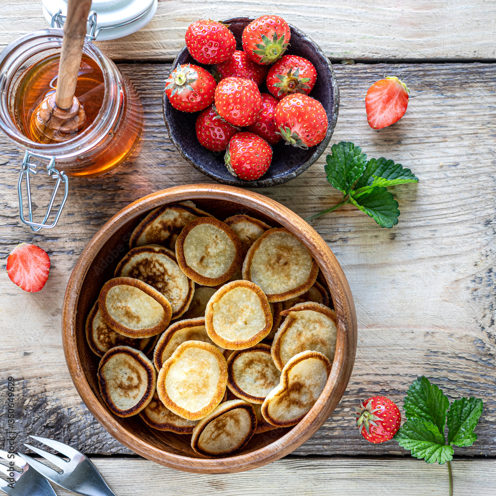Mini pancakes in a wooden bowl with strawberries and honey. Top view. Square
