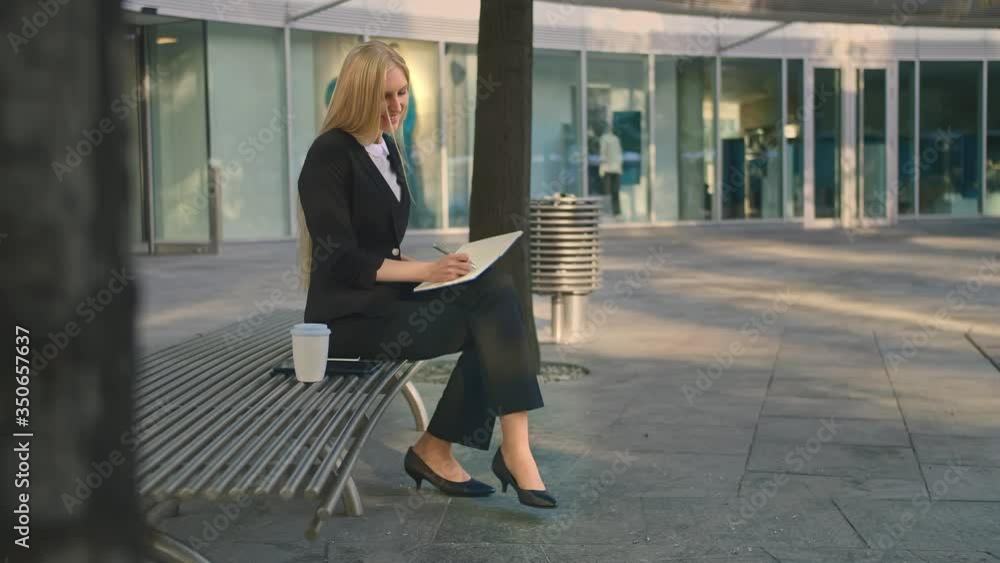 Side view of elegant businesswoman sitting on bench in patio and writing in notepad while working. Side view of elegant businesswoman sitting on bench in patio and writing in notepad while working.