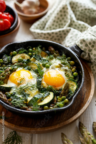 Green shakshuka with green vegetables and fried eggs in cast iron skillet on a white wooden table close up view