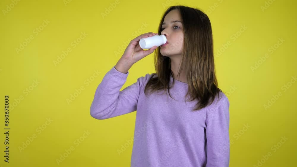Teen girl in a pink sweater drinks yogurt with a white plastic bottle for dairy products, turns to the camera, smiles and shows like on yellow background with copy space. Place for text or product Teen girl in a pink sweater drinks yogurt with a white plastic bottle for dairy products, turns to the camera, smiles and shows like on yellow background with copy space. Place for text or product
