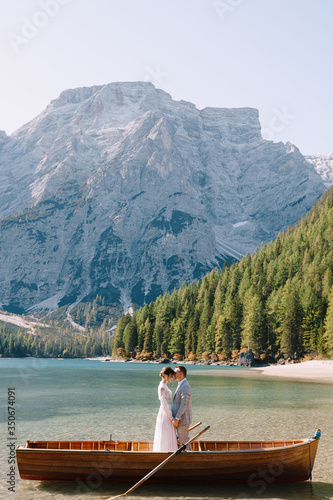 Bride and groom in a wooden boat at the Lago di Braies in Italy. Wedding couple in Europe, on Braies lake, in the Dolomites. The newlyweds are standing in the boat and hugging. © Nadtochiy