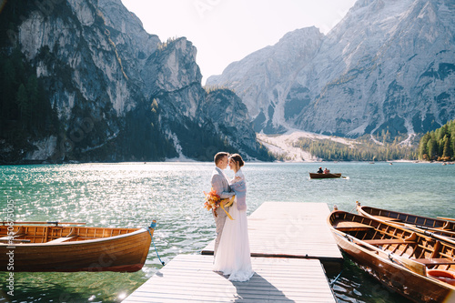 The bride and groom walk along a wooden boat dock at the Lago di Braies in Italy. Wedding in Europe, on Braies lake. Newlyweds walk, kiss, hug on a background of rocky mountains.