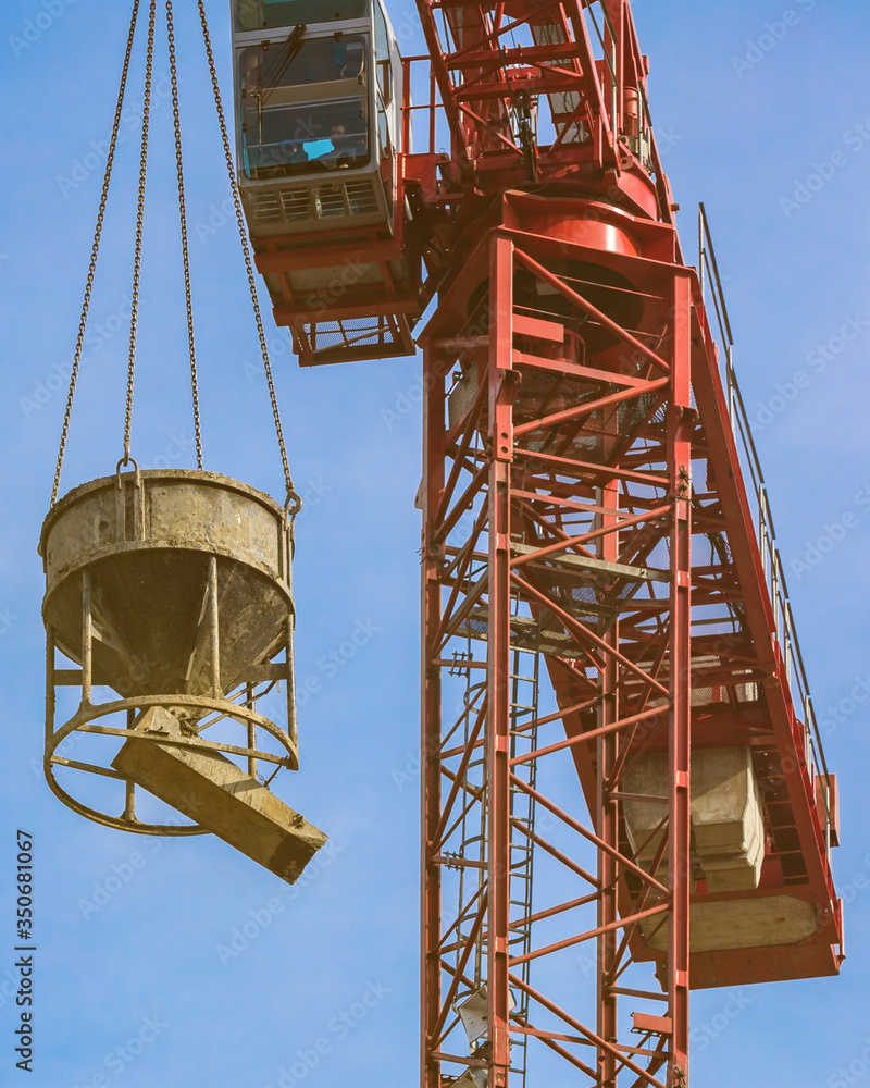 Red tower crane with cement mixer against blue sky with white clouds. Close up. Front shot. Glare of the sun on the cabin builder. Red tower crane with cement mixer against blue sky with white clouds. Close up. Front shot. Glare of the sun on the cabin builder.