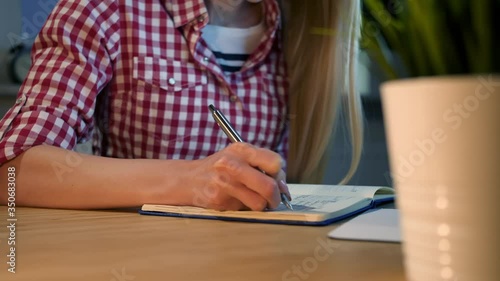 Crop view of woman with long blond hair in bright casual shirt sitting at wooden desk and writing down information with shiny metal pen into daily planner at night.