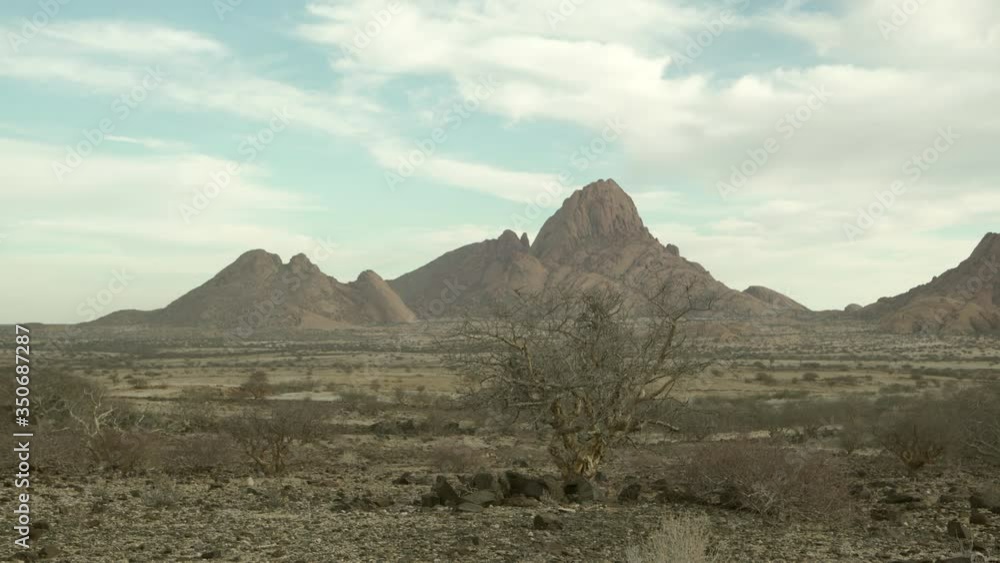 Lockdown shot of bare trees amidst rocks in landscape against cloudy sky, idyllic shot of arid desert on sunny day - Spitzkoppe, Namibia Lockdown shot of bare trees amidst rocks in landscape against cloudy sky, idyllic shot of arid desert on sunny day - Spitzkoppe, Namibia