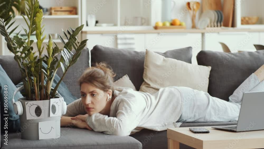 Young positive woman sitting on couch, holding smartphone, waving and talking about her houseplant in pot decorated with smiley face while video calling from home during quarantine Young positive woman sitting on couch, holding smartphone, waving and talking about her houseplant in pot decorated with smiley face while video calling from home during quarantine
