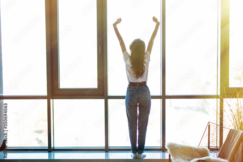 Good morning. Back view of happy young woman stretching and looking out the window at home Good morning. Back view of happy young woman stretching and looking out the window at home