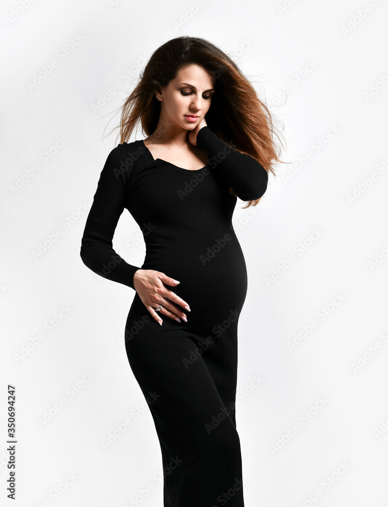 Brunette pregnant woman in black tight dress. She put hand on her belly while posing sideways isolated on white. Close up Brunette pregnant woman in black tight dress. She put hand on her belly while posing sideways isolated on white. Close up