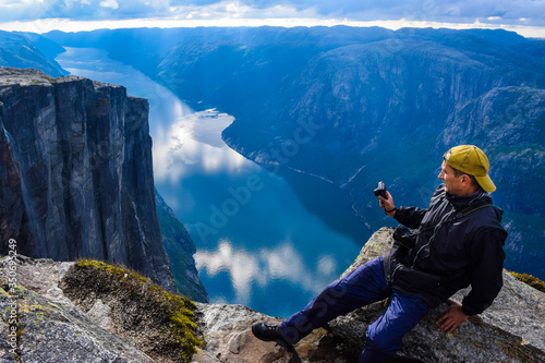 Tourist man on the edge above Lysefjorden. Wonderful mountain landscape with clouds reflected in blue water, where Kjeragbolten is located nearby. View from Kjerag plateau, Norway.