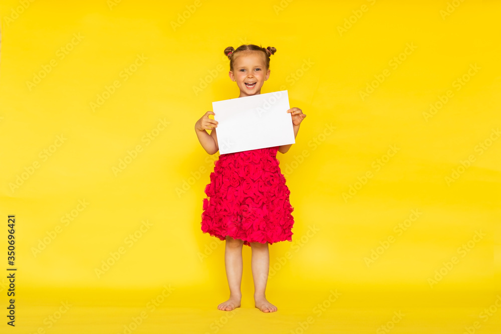 Pretty little barefooted girl with two buns and in bright pink summer dress posing with white paper list on yellow background. Copy space Pretty little barefooted girl with two buns and in bright pink summer dress posing with white paper list on yellow background. Copy space