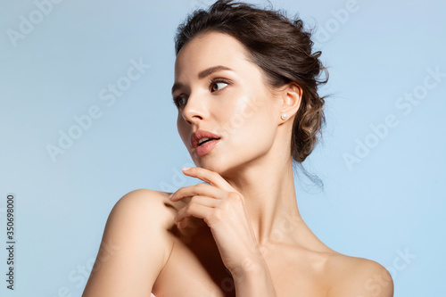 Beauty portrait of young woman with shiny skin standing on blue copy space and looking aside touching chin with hand.