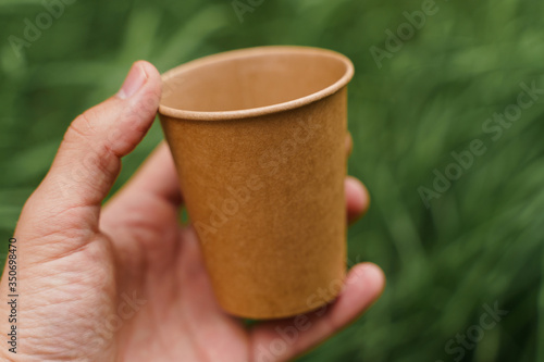 Close up isolated eco-friendly natural coffee cup. Disposable ecological utensils in hands on green grass background. Sustainability of planet. Cardboard glass made of fiber of bamboo and bagasse