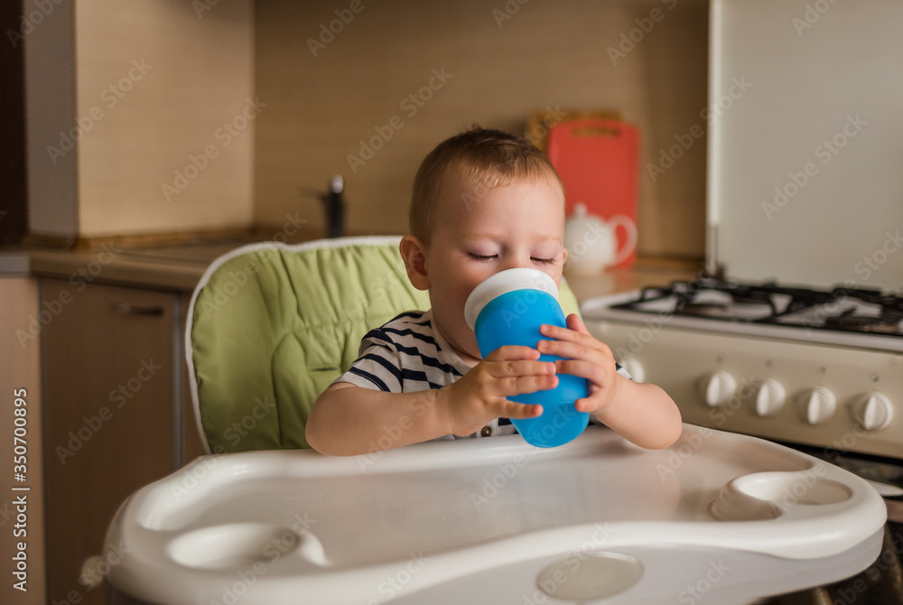 A small boy in a striped t-shirt sits on a high chair and drinks water from a bottle. A small boy in a striped t-shirt sits on a high chair and drinks water from a bottle.
