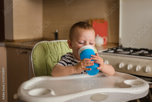 A small boy in a striped t-shirt sits on a high chair and drinks water from a bottle.