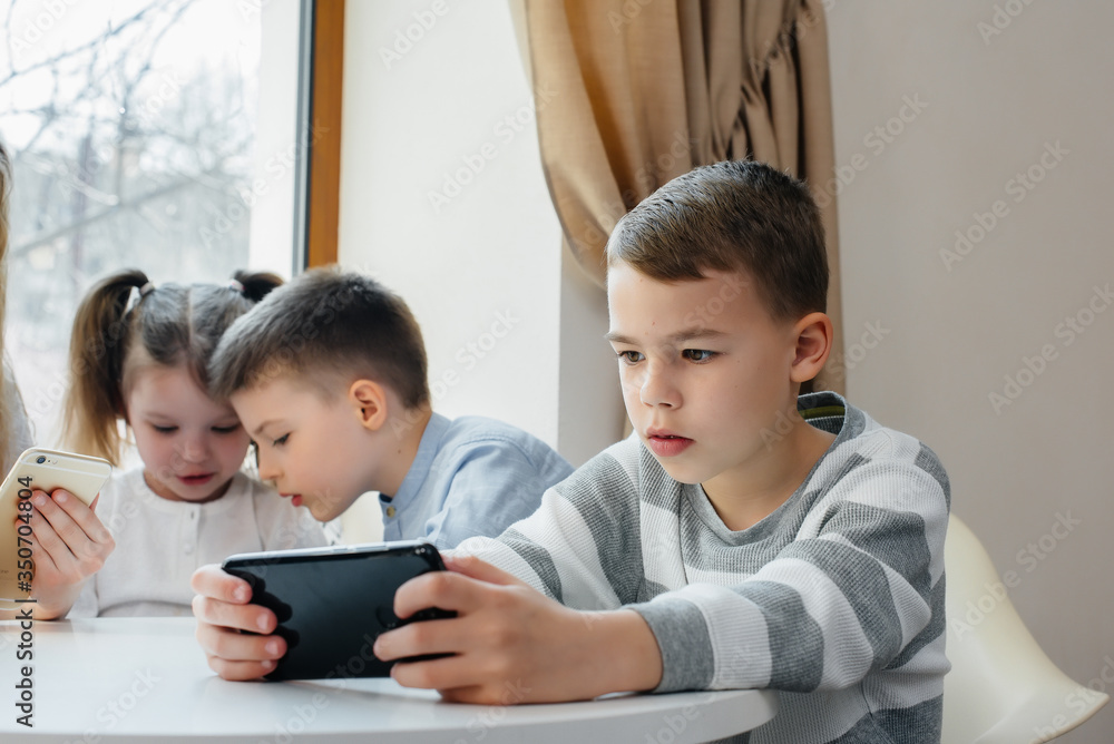 Children sit at a table in a cafe and play mobile phones together. Modern entertainment Children sit at a table in a cafe and play mobile phones together. Modern entertainment
