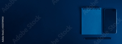 notebook on a spring, and a pen, and mobile phone on a black background. space for text. banner. toned classic blue color