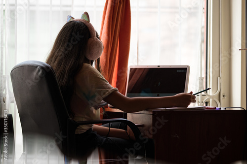 Young woman working at home with computer, notebook, pencils and pens at her desk; she indoors. She's wearing headphones with cat's ears and is beside the window. Bogota, Colombia.