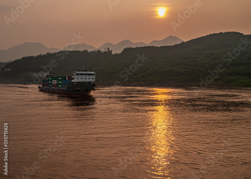 Chongqing, China - May 8, 2010: Evening light on Yangtze River. Container ship sails on while golden sunset reflected by brown water in front of shoreline with sun above forested hills.