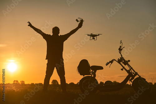 The flying drone pilot controls the drone in the rays of the setting sun. Nearby is an electric scooter.