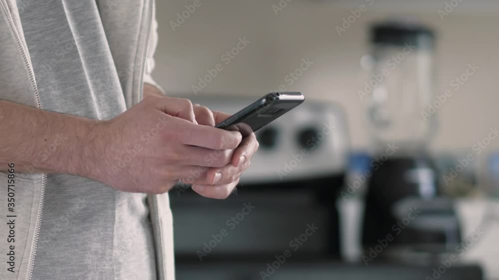 Closeup of a man standing in his kitchen at home and typing on his smartphone mobile device. Closeup of a man standing in his kitchen at home and typing on his smartphone mobile device.