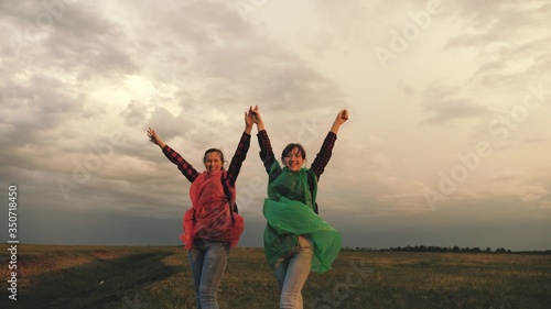 strong and healthy girls travelers travel in colorful raincoats, climb the mountain, hold hands, help each other. Free women tourists with backpacks on rise to top of hill, rejoice in victory and jump
