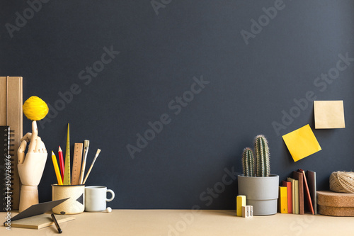 Creative desk with a blank picture frame or poster, desk objects, office supplies, books, and plant on a dark blue background. 