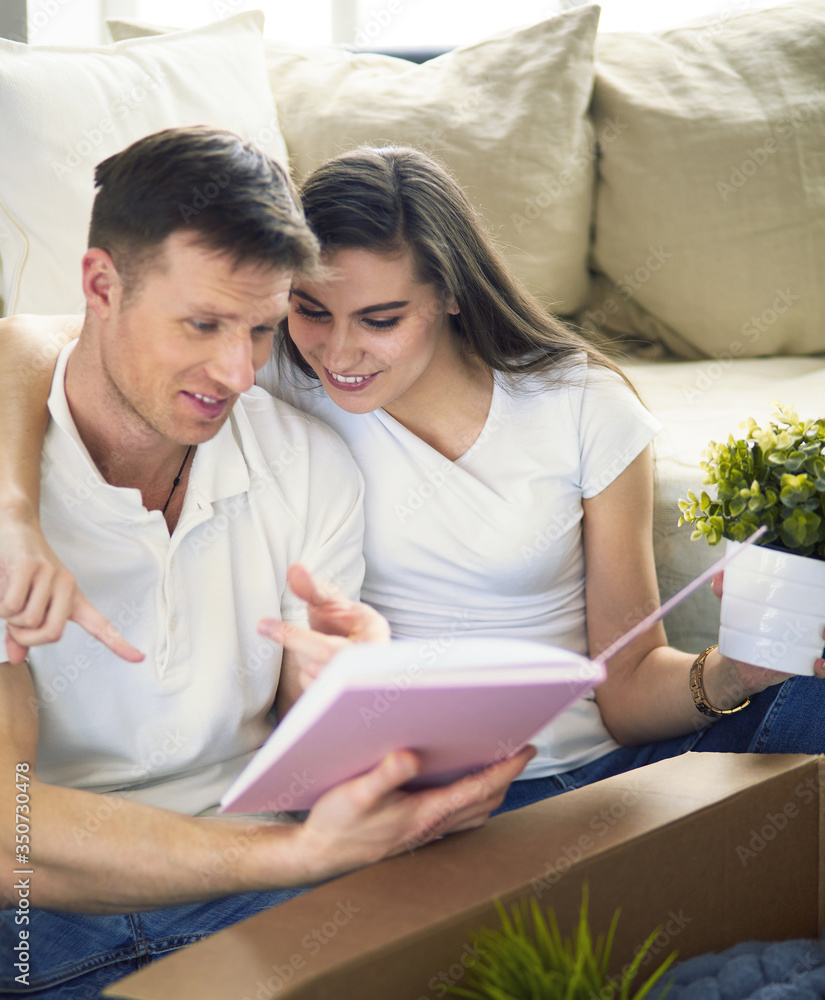 Cute couple unpacking cardboard boxes in their new home, sitting on the floor and looking at a family album Cute couple unpacking cardboard boxes in their new home, sitting on the floor and looking at a family album