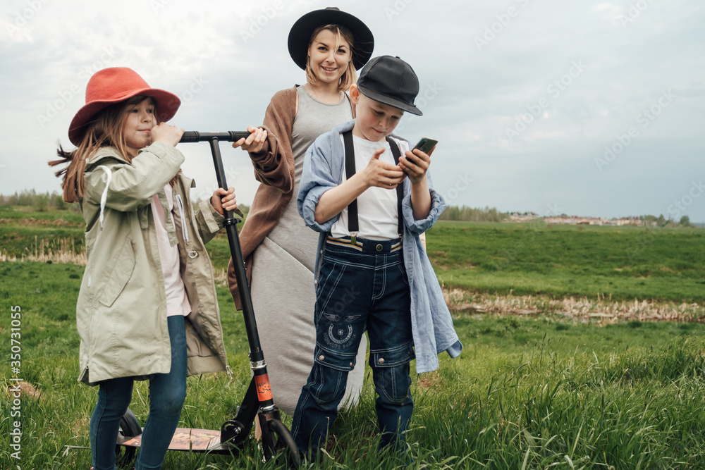Happy family, mother, daughter and son in organic clothes, walk in the field and ride a scooter and watch video on the phone. The concept of a healthy lifestyle, happiness and joy