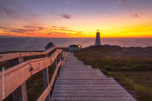 Konstfotografi Wooden staircase and boardwalk at a beautiful sunrise, overlooking a white lighthouse sitting at the edge of a rocky cliff